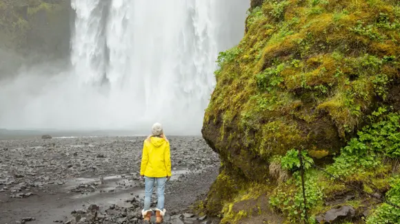 A female tourist in a yellow jacket standing before a waterfall in Iceland.