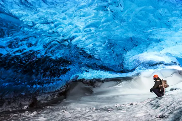 An explorer crouches on a snowy path, marvelling at the ethereal blue glow of an ice cave's arching ceiling