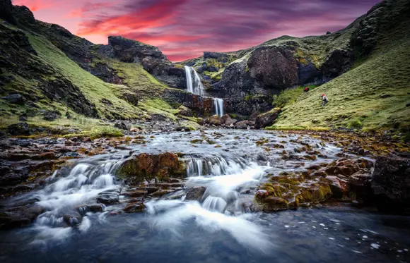 A serene waterfall in Iceland, with a stream in the foreground and a dramatic sky with hues of pink and blue